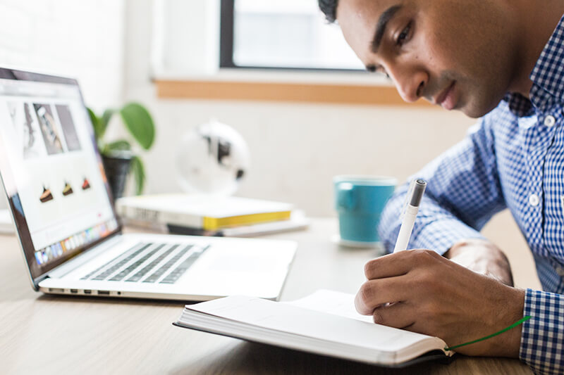 Male student writing on paper in front of a laptop