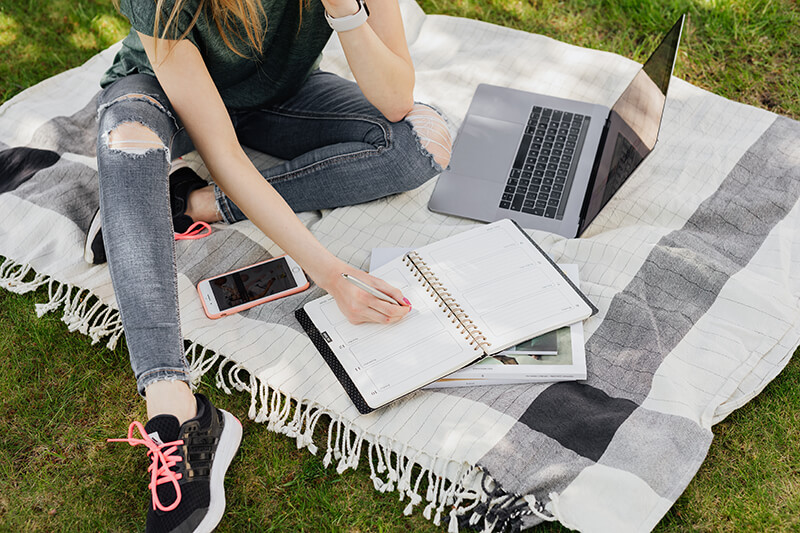 College student sitting on a picnic blanket in a park working on her descriptive essay