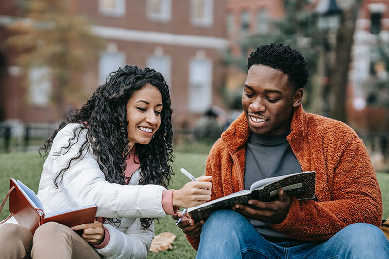 Two students sitting in the grass offering essay writing help to each other