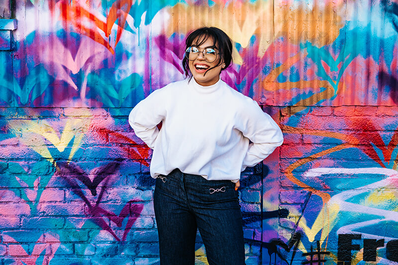 woman smiling as she poses in front of a colorful mural