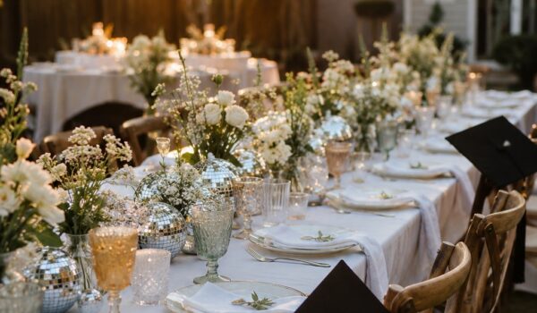An outdoor college graduation party table set with florals, hanging disco balls, string lights, and place settings at golden hour